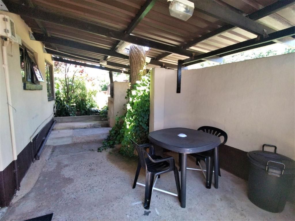 A covered patio with a round table and four chairs, a trash bin, and a staircase leading to an upper level.