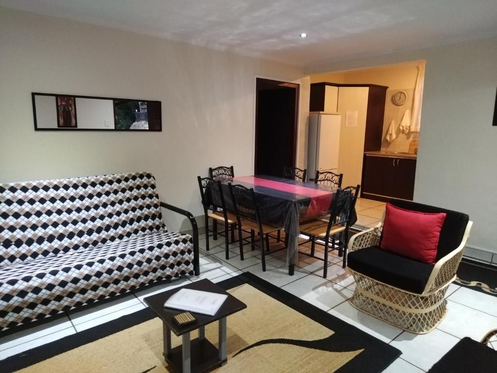A living room with a sofa, a dining table with chairs, a wicker chair, and a kitchen area in the background.