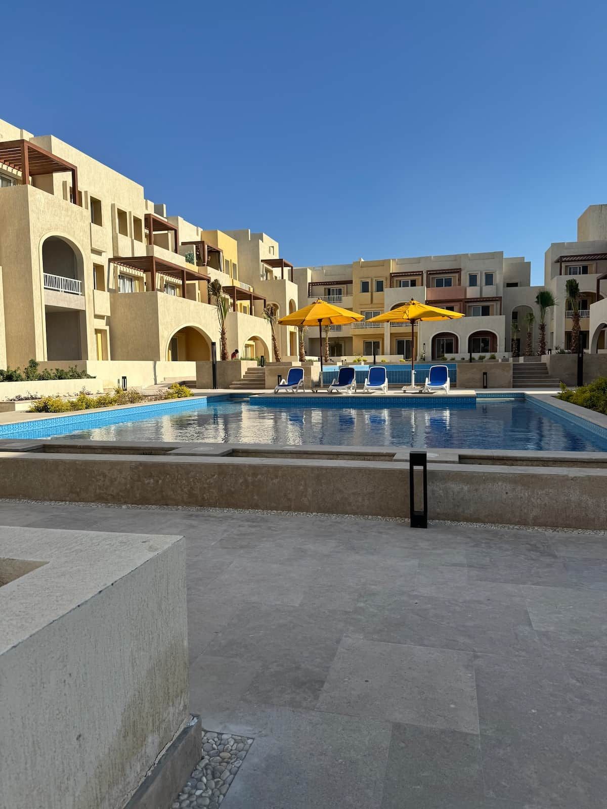 Una piscina con sillas de descanso y sombrillas, rodeada de edificios de varios pisos con balcones.