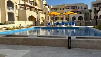 Una piscina con sillas de descanso y sombrillas, rodeada de edificios de varios pisos con balcones.