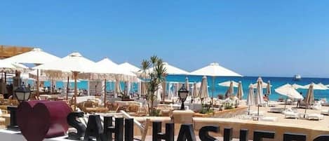 A beachside resort with sun loungers and umbrellas, a clear blue sky, and a sign reading 'SAHL HASHEES'.