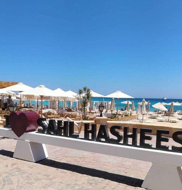 Ein Strandresort mit Liegestühlen und Sonnenschirmen, einem klaren blauen Himmel und einem Schild mit der Aufschrift 'SAHL HASHEES'.