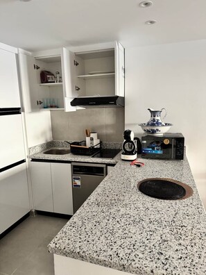 Eine kompakte Küche mit weißen Schränken, einer Granitarbeitsplatte, einem Geschirrspüler, einer Kaffeemaschine und einer Mikrowelle.