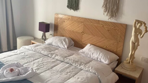 Cama bem arrumada com cabeceira de madeira, mesa de cabeceira com abajur e um detalhe de macramé na parede.