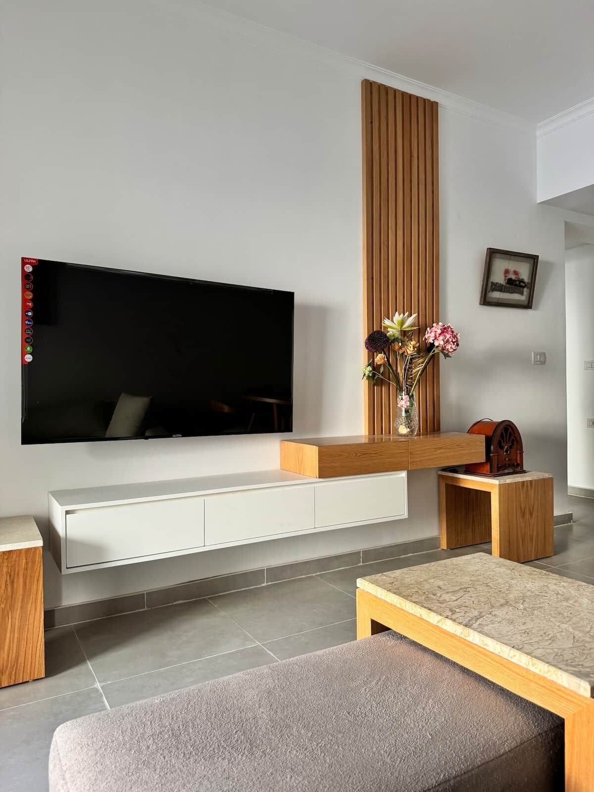 Ein modernes Wohnzimmer mit einem Flachbildfernseher an einer weißen Konsole, einer Holz-Akzentwand, einer Vase mit Blumen und einem kleinen Holztisch.