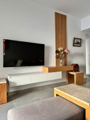 Een moderne woonkamer met een flatscreen televisie bevestigd aan een witte console, een houten accentmuur, een vaas met bloemen en een kleine houten tafel.