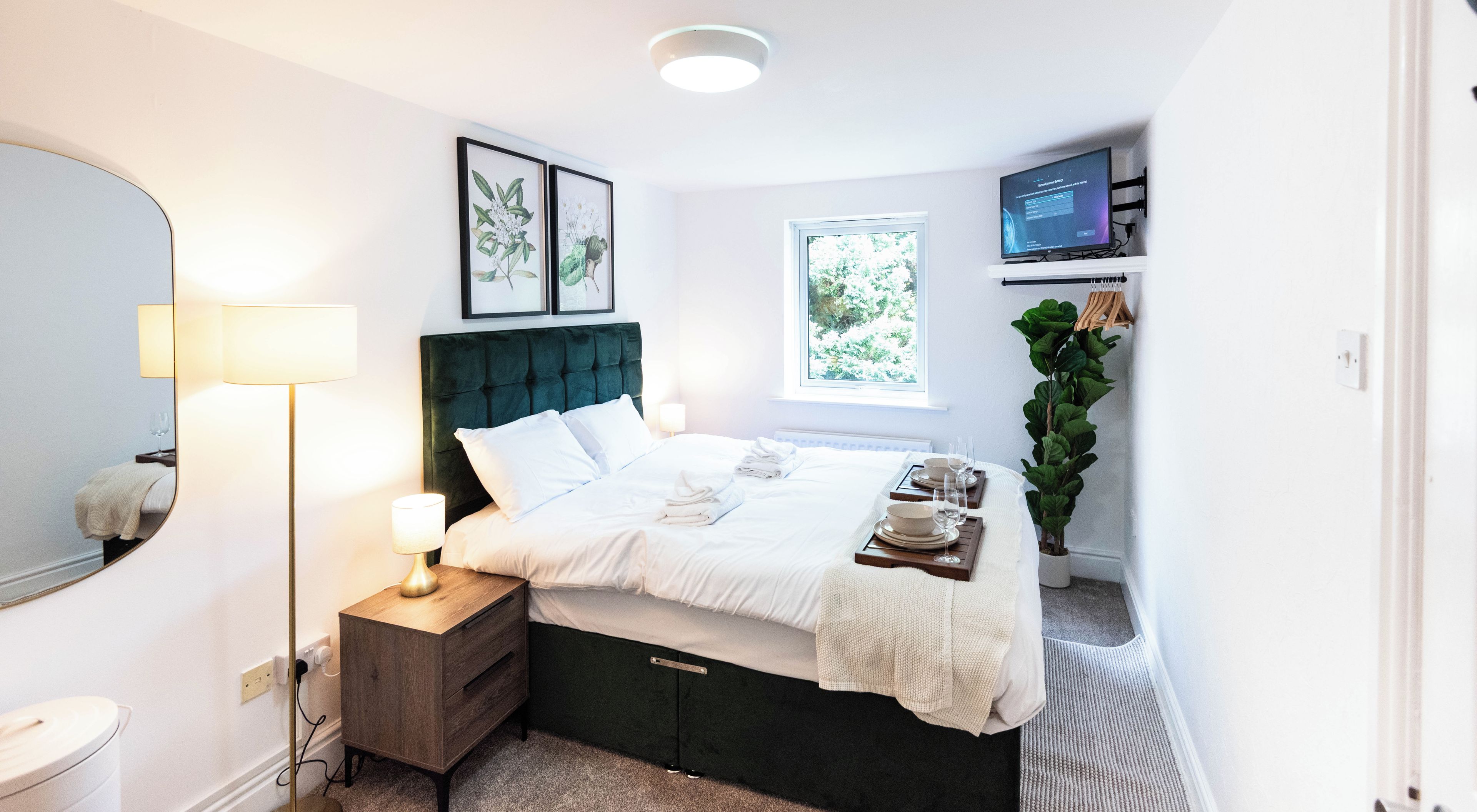 A bedroom with a bed, bedside table, lamp, artwork, window, and a television.