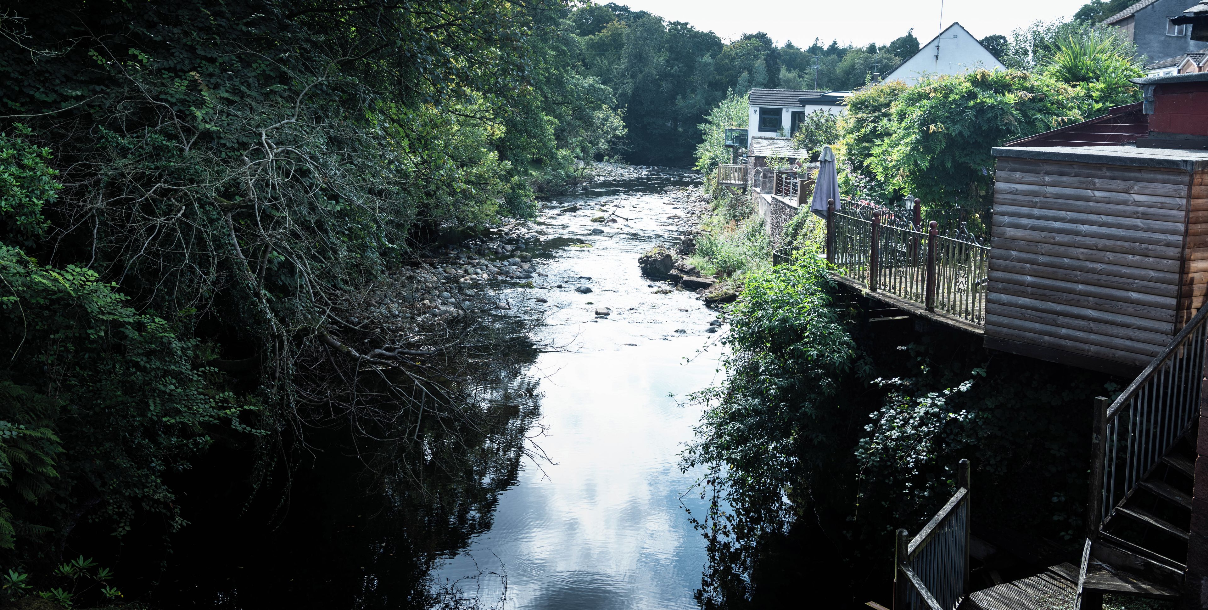 A river flowing through a wooded area with a wooden bridge and buildings on the right.