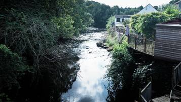 A river flowing through a wooded area with a wooden bridge and buildings on the right.