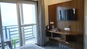 A hotel room with a bed, a TV mounted on a wooden panel, a small table with a kettle and glasses, and a balcony with a green chair.
