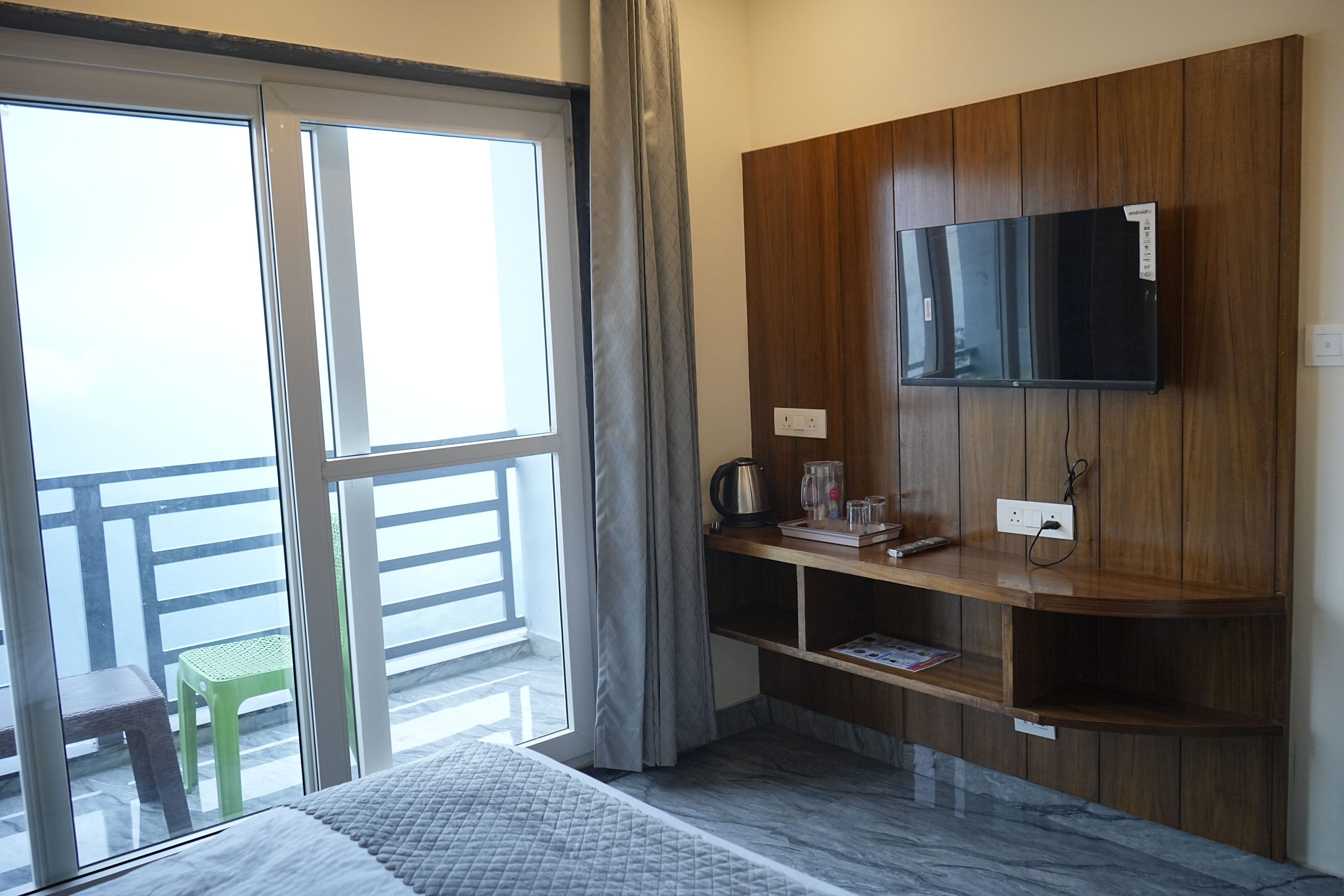 Quarto de hotel com cama, TV fixada em painel de madeira, mesinha com chaleira e copos, e vista para o exterior por uma grande janela.