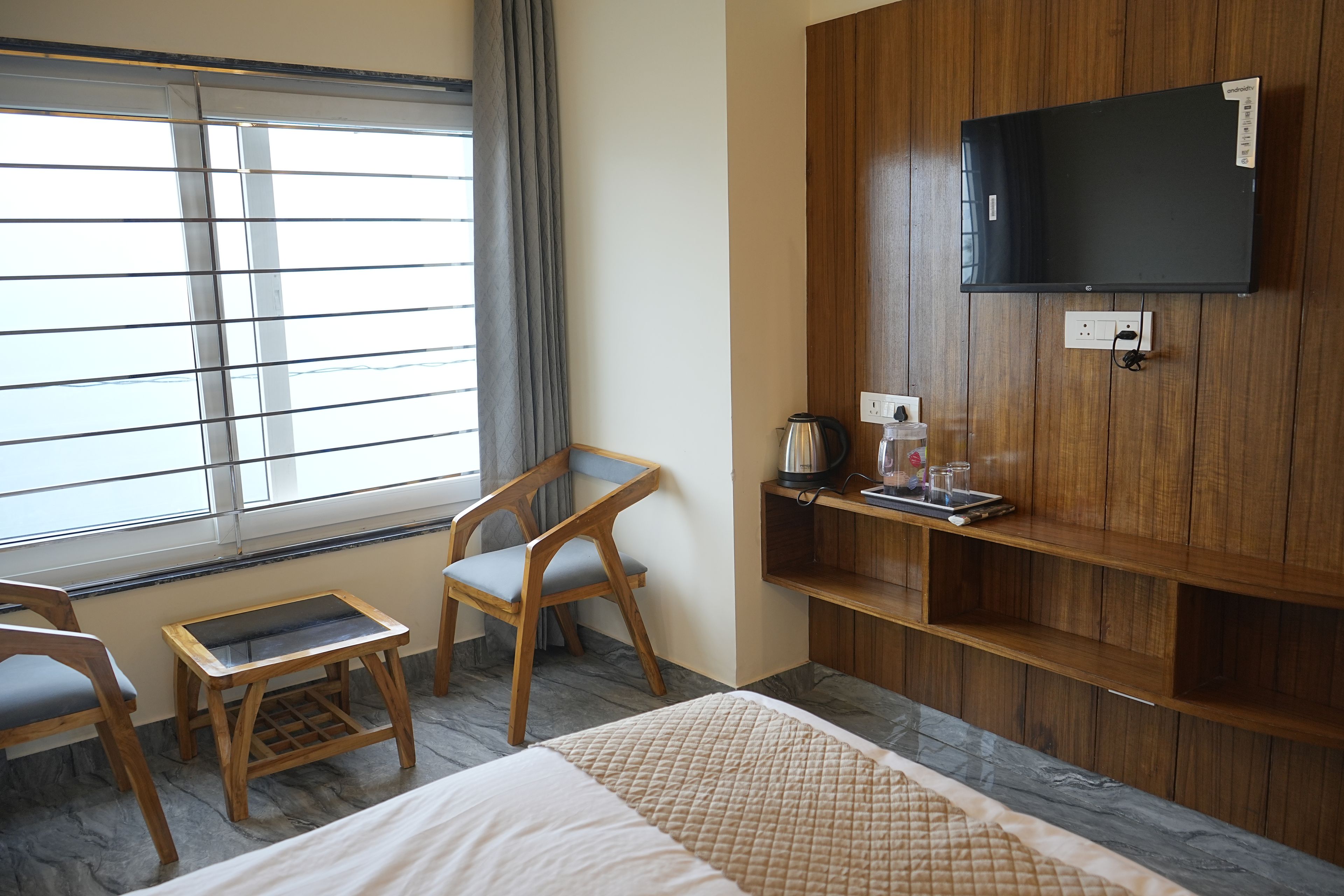 A hotel room with a bed, a TV mounted on the wall, a window with blinds, a chair, and a small table with a kettle and glasses.