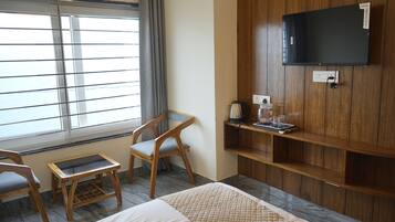 A hotel room with a bed, a TV mounted on the wall, a window with blinds, a chair, and a small table with a kettle and glasses.