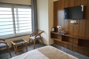 A hotel room with a bed, a TV mounted on the wall, a window with blinds, a chair, and a small table with a kettle and glasses. - The Vanaaya luxury Homestay Mussoorie (Mussoorie)