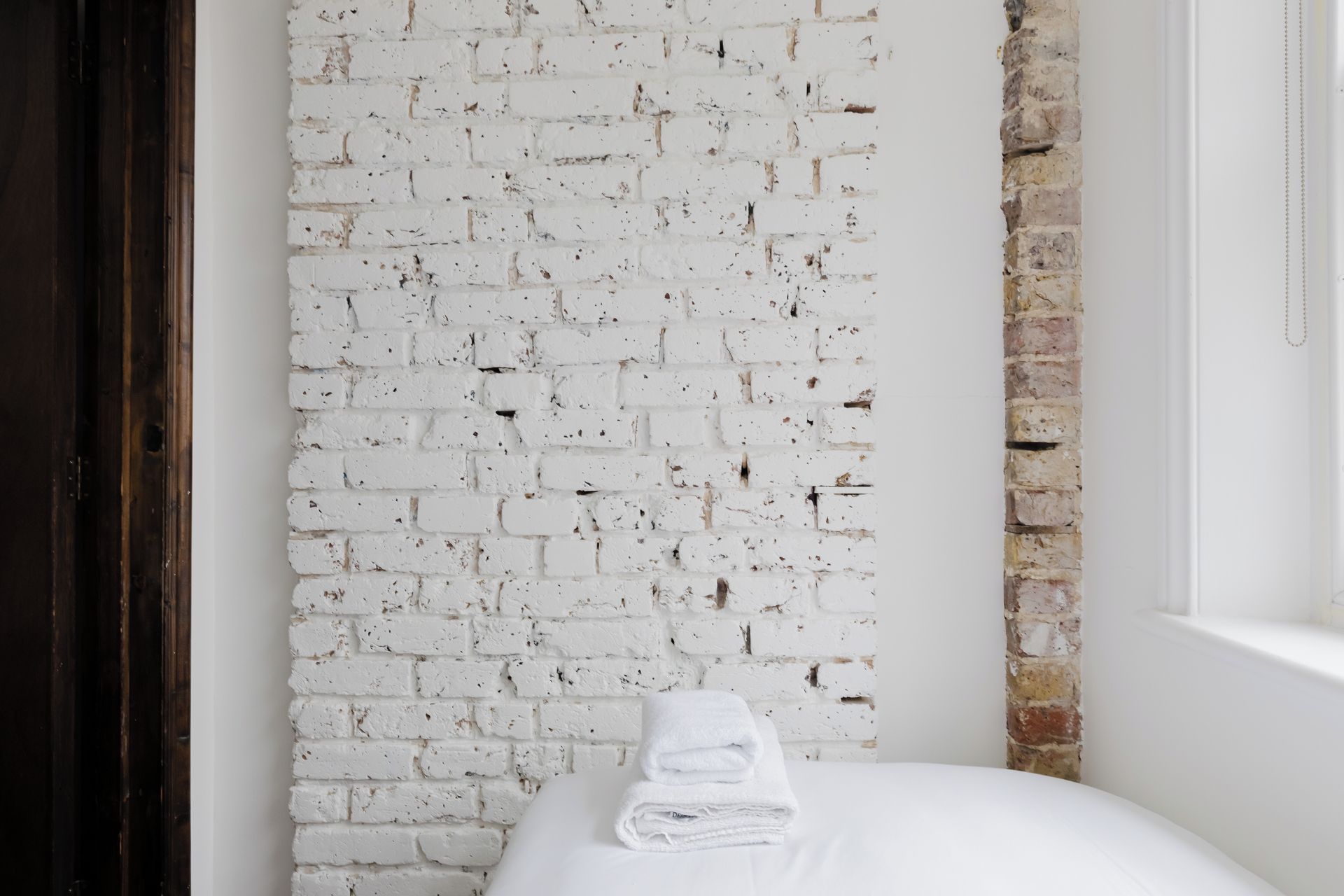 A room with a white brick wall, a wooden door, and a white bed with a folded white towel.