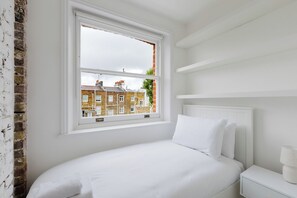 Ein Schlafzimmer mit einem weißen Bett, einem Nachttisch und einem Fenster mit Blick auf Ziegelbauten.