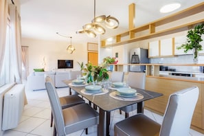A modern dining area with a table set for four, a kitchen in the background, and a living area with a sofa and TV. - Plumeria Apartment Chania (Theriso)