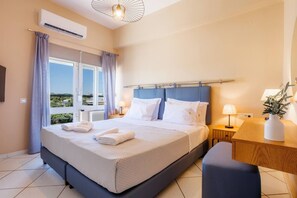 A hotel room with a bed, a nightstand, a desk, a chair, a window with curtains, and an air conditioning unit. - Plumeria Apartment Chania (Theriso)
