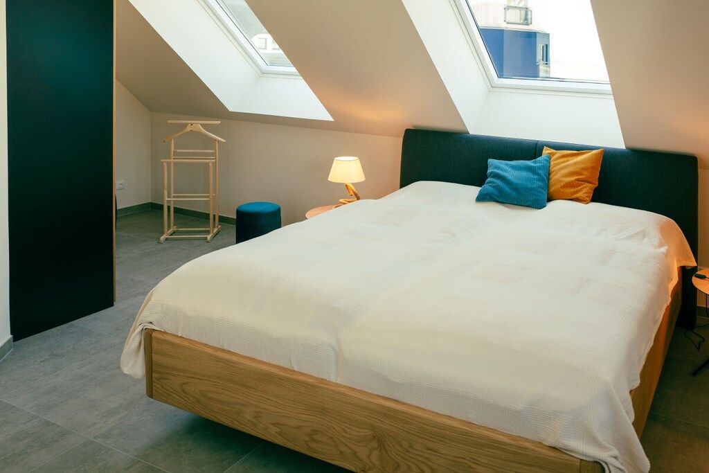 Un dormitorio con tragaluces, una cama con colchón blanco, una mesita de noche con lámpara y un perchero.