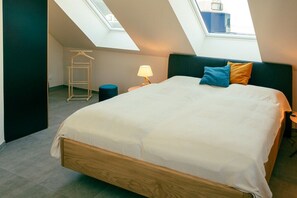Quarto com claraboia, cama com colchão branco, mesa de cabeceira com abajur e um cabideiro.