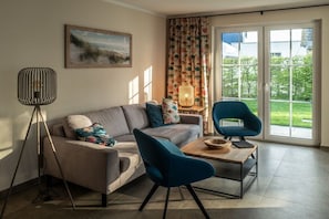 A living room with a sofa, a blue armchair, a floor lamp, a table, and a window with a view of greenery. - Holiday home with 3 bedrooms (Breege\/ Juliusruh OT Breege)