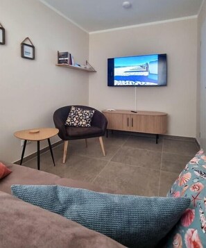 Sala de estar com televisão de tela plana na parede, poltrona escura com almofada estampada, mesa redonda pequena e sofá com almofada azul.