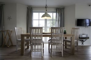 A dining area with a wooden table and chairs, a hanging light fixture, a television on the wall, and a window with curtains. - Holiday home with 2 bedrooms (Fuhlendorf)