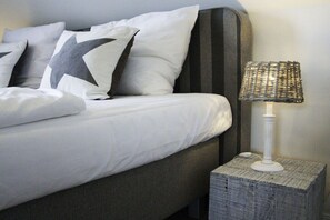 Una cama bien hecha con un almohadón grande con forma de estrella, una lámpara blanca sobre una mesita de noche con textura, y un cabecero oscuro.