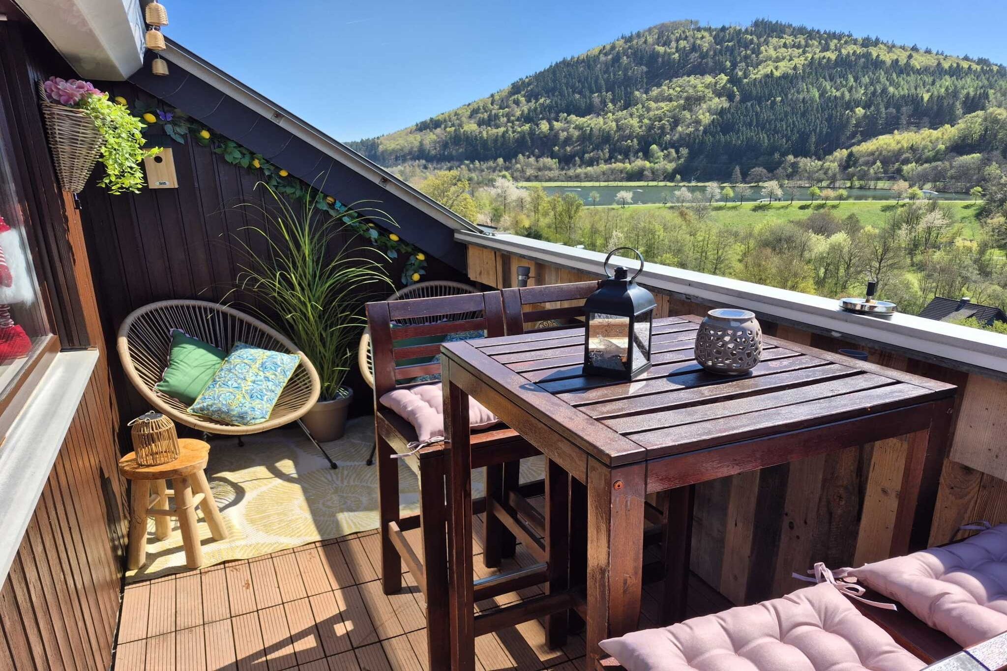 Eine Holterrasse mit Tisch und Stühlen, eine hängende Pflanze und Blick auf ein grünes Tal.