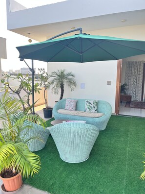 A rooftop terrace with a green artificial grass area, a wicker sofa set, and a large umbrella providing shade. - Cozy 1-bedroom on a rooftop in a bungalow Jaipur with terrace garden (Jaipur)