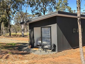 A modern building with a large glass door, a small patio with a table and chairs, surrounded by trees. - Villa 1 @ North Peak - A one bedroom villa for two (Pokolbin)