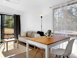A modern living room with a wooden table, white sofa, and a potted plant. - Villa 1 @ North Peak - A one bedroom villa for two (Pokolbin)