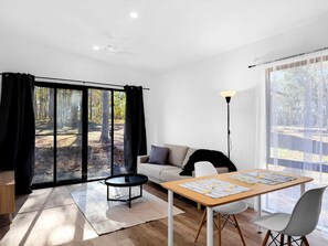 Ein modernes Wohnzimmer mit einer Couch, einem runden Couchtisch, einem Esstisch mit St ühlen, einer Stehlampe und großen Fenstern mit Vorhängen.