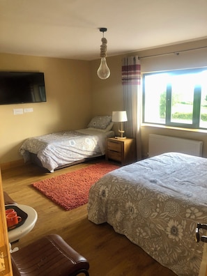 A bedroom with two beds, a nightstand with a lamp, a red rug, a wooden bench, and a TV mounted on the wall.
