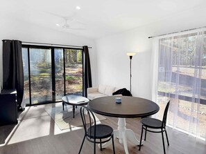 A modern room with a round dining table, two chairs, a sofa, and a large window with a view of a forest. - Villa 3 @ North Peak - A cute cabin for two (Pokolbin)
