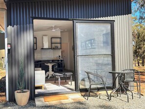 A modern outdoor seating area with a table and chairs, a potted plant, and a sliding glass door leading to a kitchen. - Villa 3 @ North Peak - A cute cabin for two (Pokolbin)