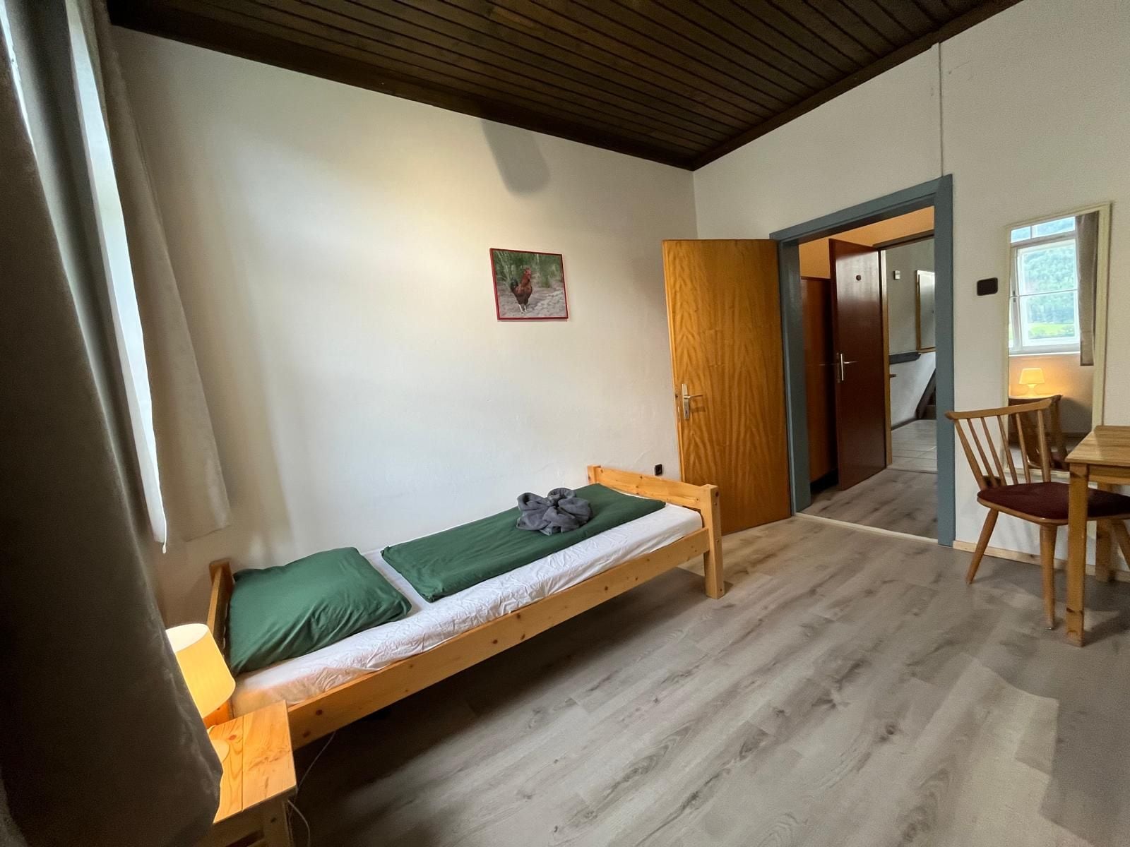 Habitación individual con una cama de madera, un edredón verde, una mesita con una silla y una puerta que da a otra habitación.