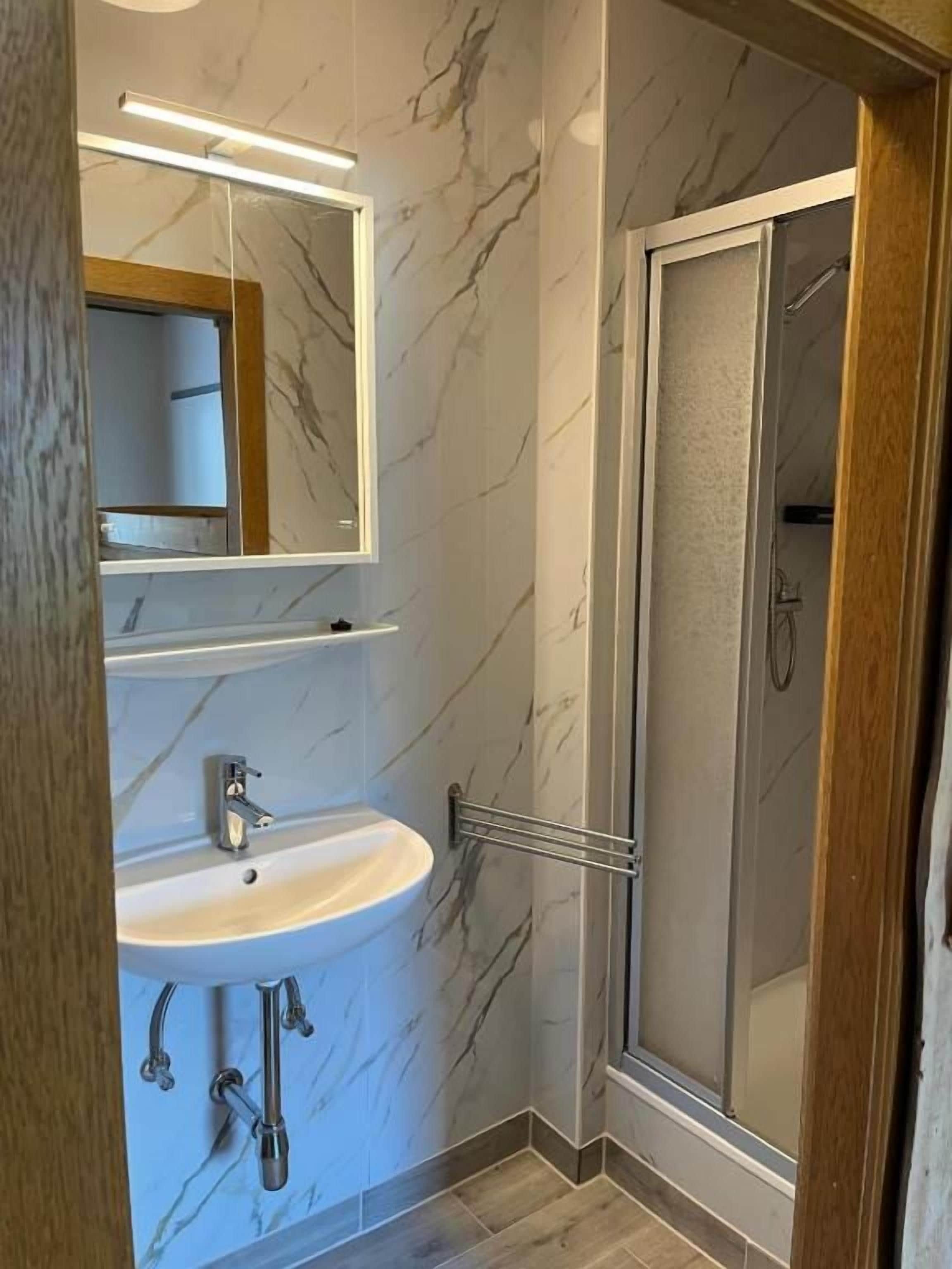 A modern bathroom with a marble wall, a white sink, a mirror, and a glass-enclosed shower.