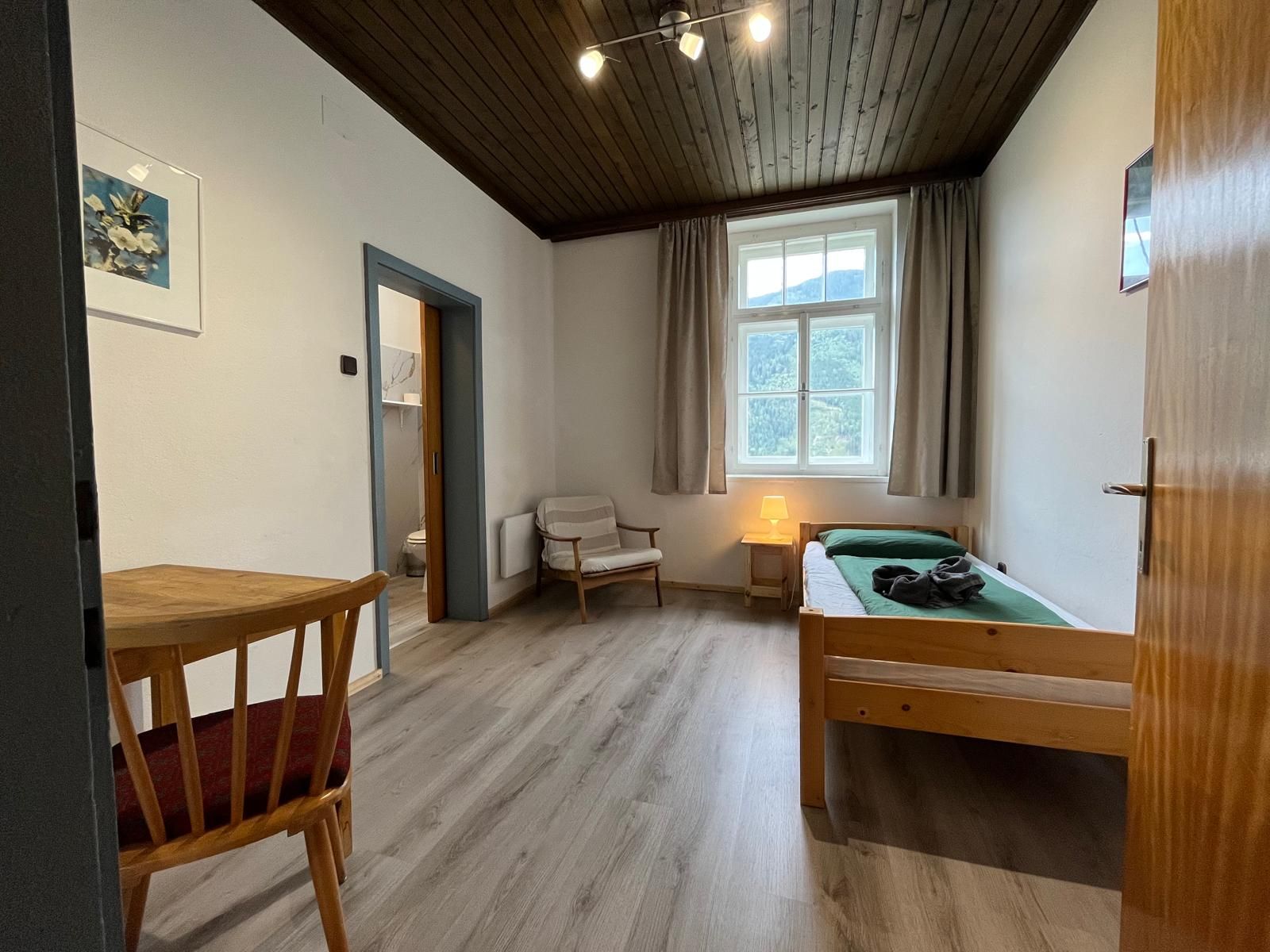 Une pièce avec un plancher en bois, un lit recouvert d’une couverture verte, une chaise, une petite table, une fenêtre avec des rideaux et un luminaire au plafond.