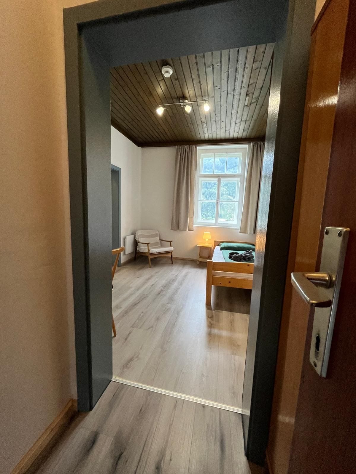 Et værelse med en seng, en stol, et vindue med gardiner samt et loft med træbjælker og lamper.