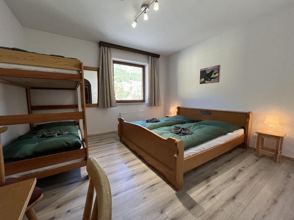 A room with bunk beds, a desk, a chair, a bedside table with a lamp, and a window with curtains.