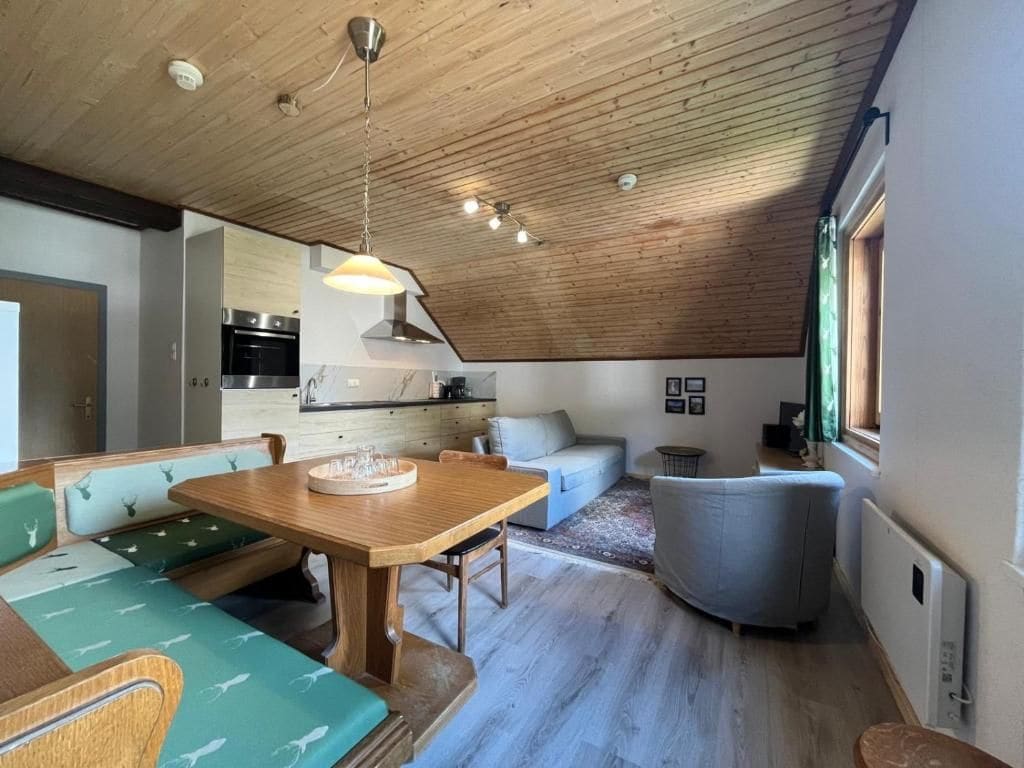 A modern interior with a wooden ceiling, a dining area with a wooden table and bench seating, a kitchen with stainless steel appliances, and a gray sofa.