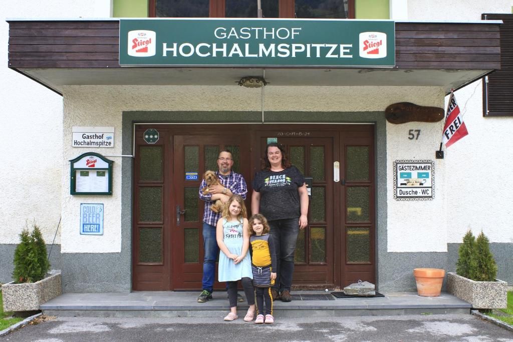 En familie står foran en bygning, hvor der står 'GASTHOF HOCHALMSPITZE' på skiltet.