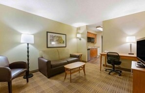 A hotel room with a sofa, a small table, a desk with a chair, a television, and a kitchenette.