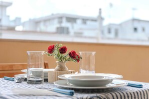 A dining table with plates, glasses, and cutlery, a vase with red flowers, and a view of white buildings in the background. - 100 meters from the beach (Alghero)