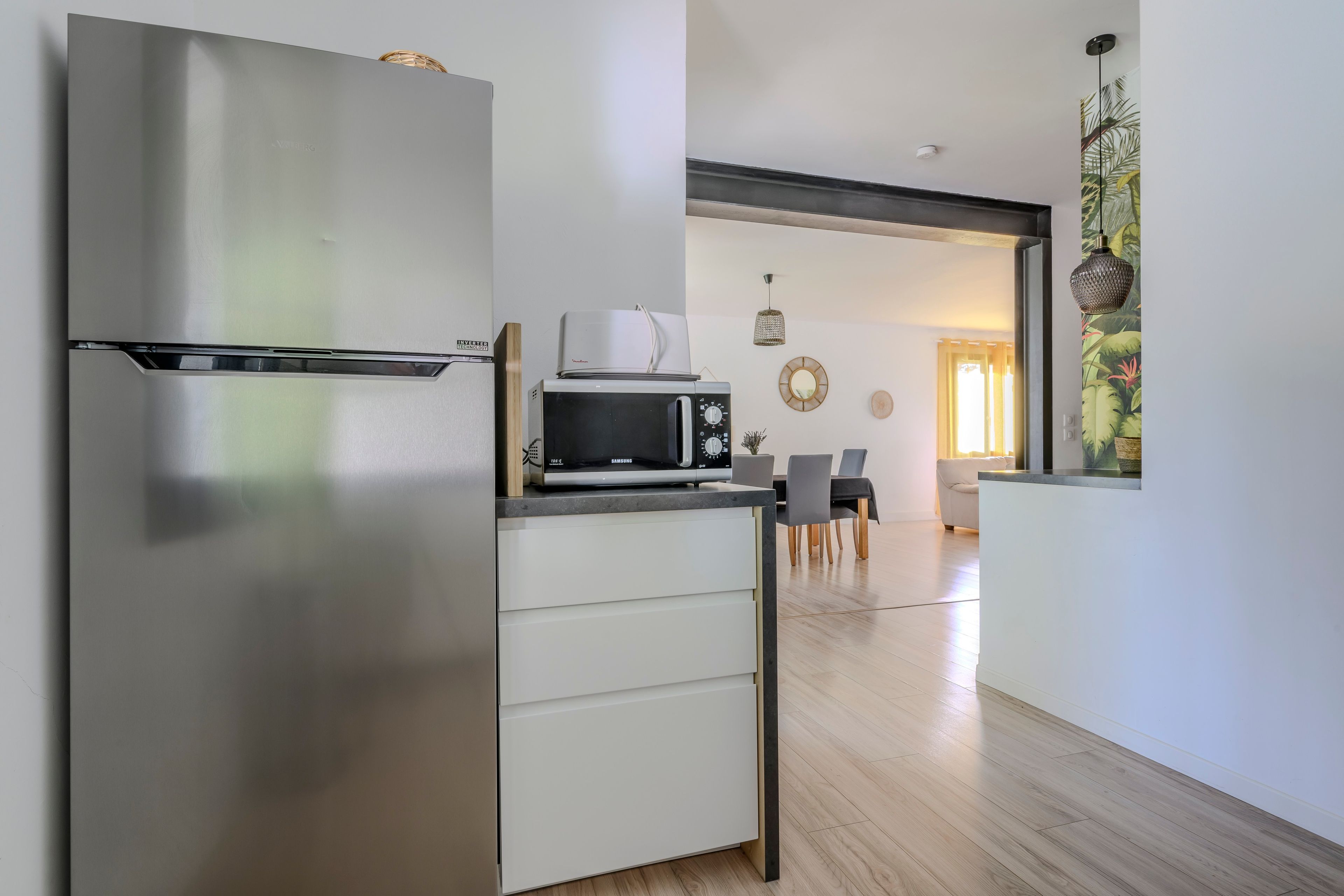 A modern kitchen with a stainless steel refrigerator, a microwave, and a built-in oven. There is a dining area with a table and chairs visible in the background.