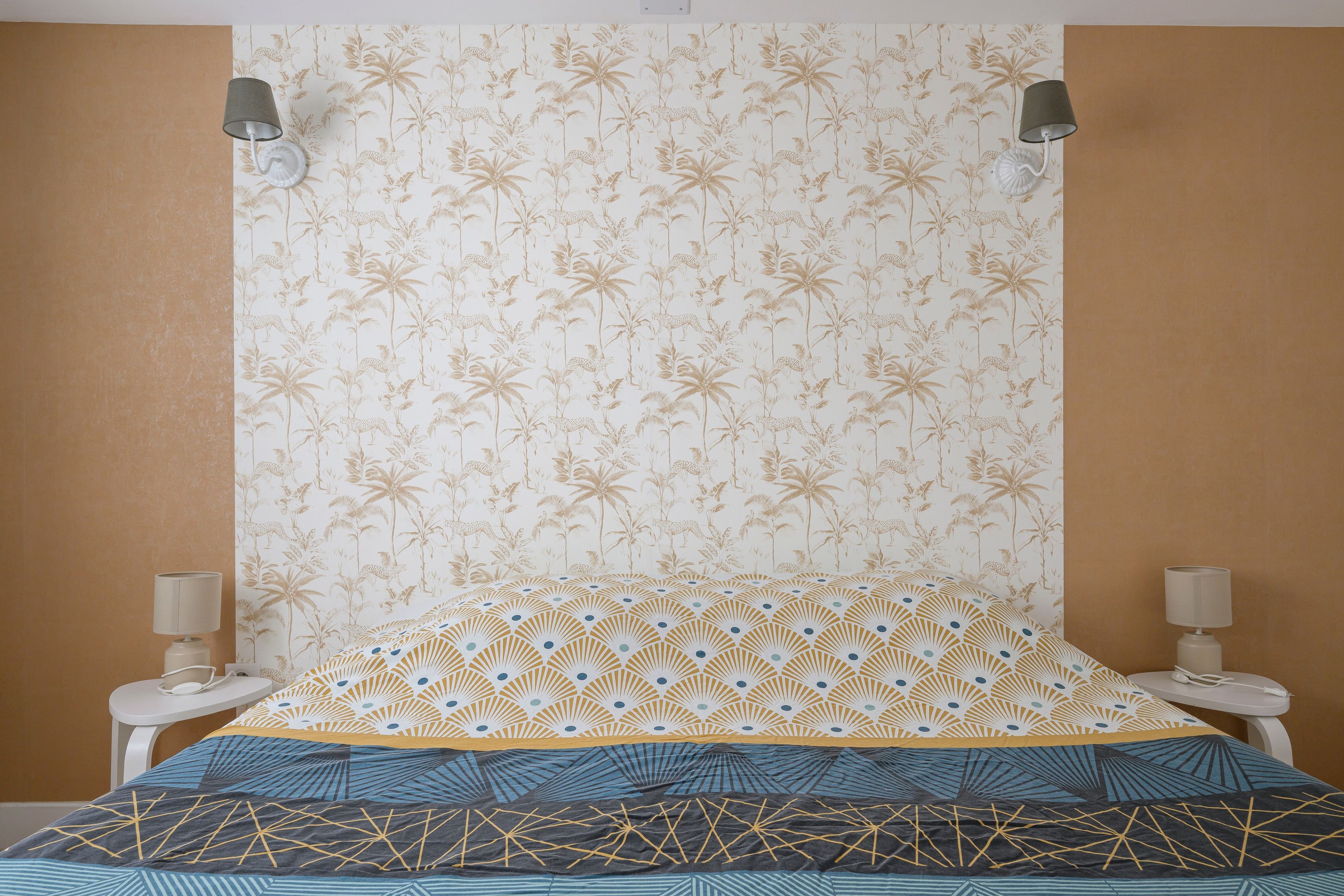Une chambre avec un papier peint à motifs, un lit recouvert d’une couvre-lit à motifs géométriques, deux lampes de chevet et une table de chevet blanche.