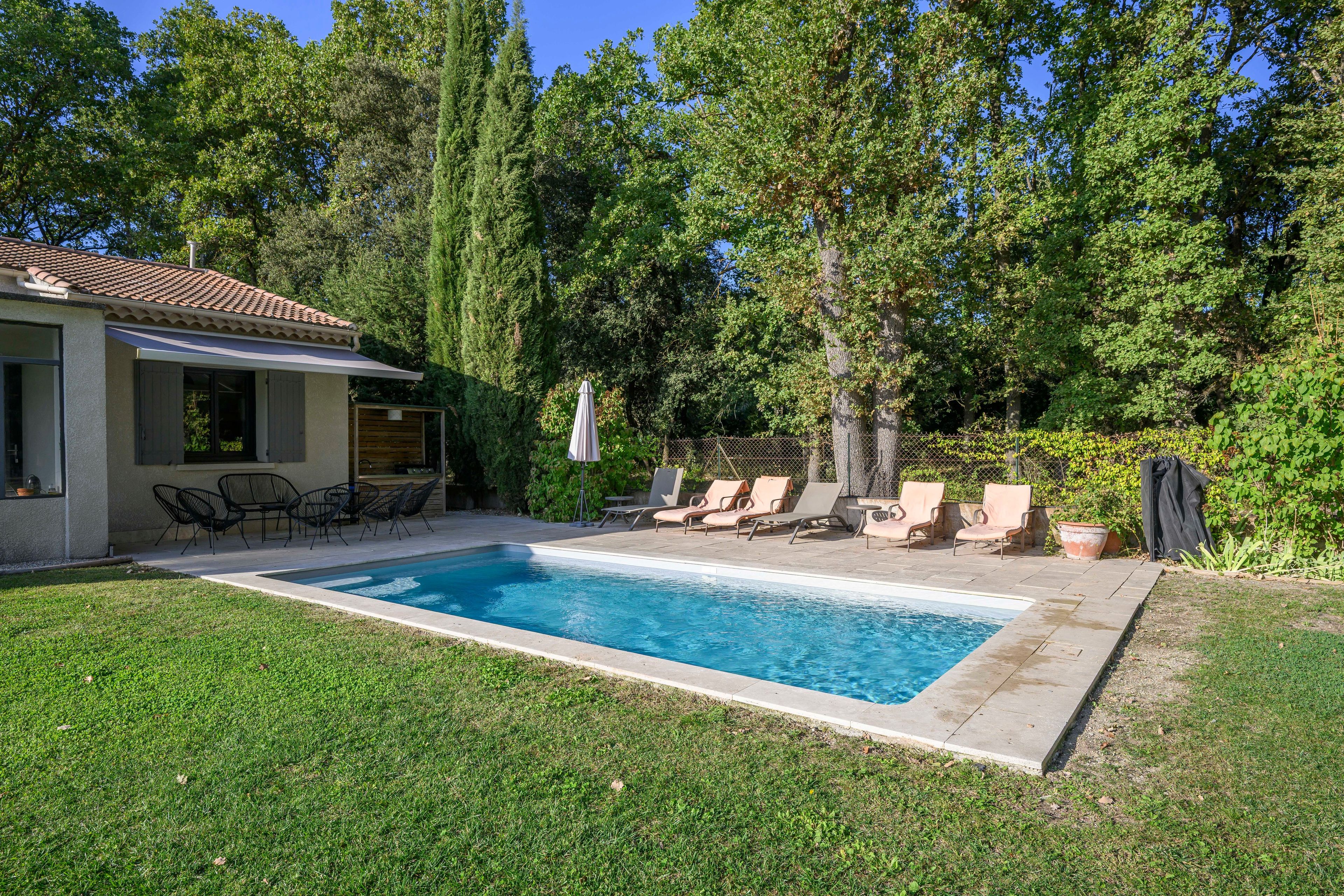 A rectangular swimming pool with lounge chairs and a patio table, surrounded by a well-maintained lawn and tall trees.