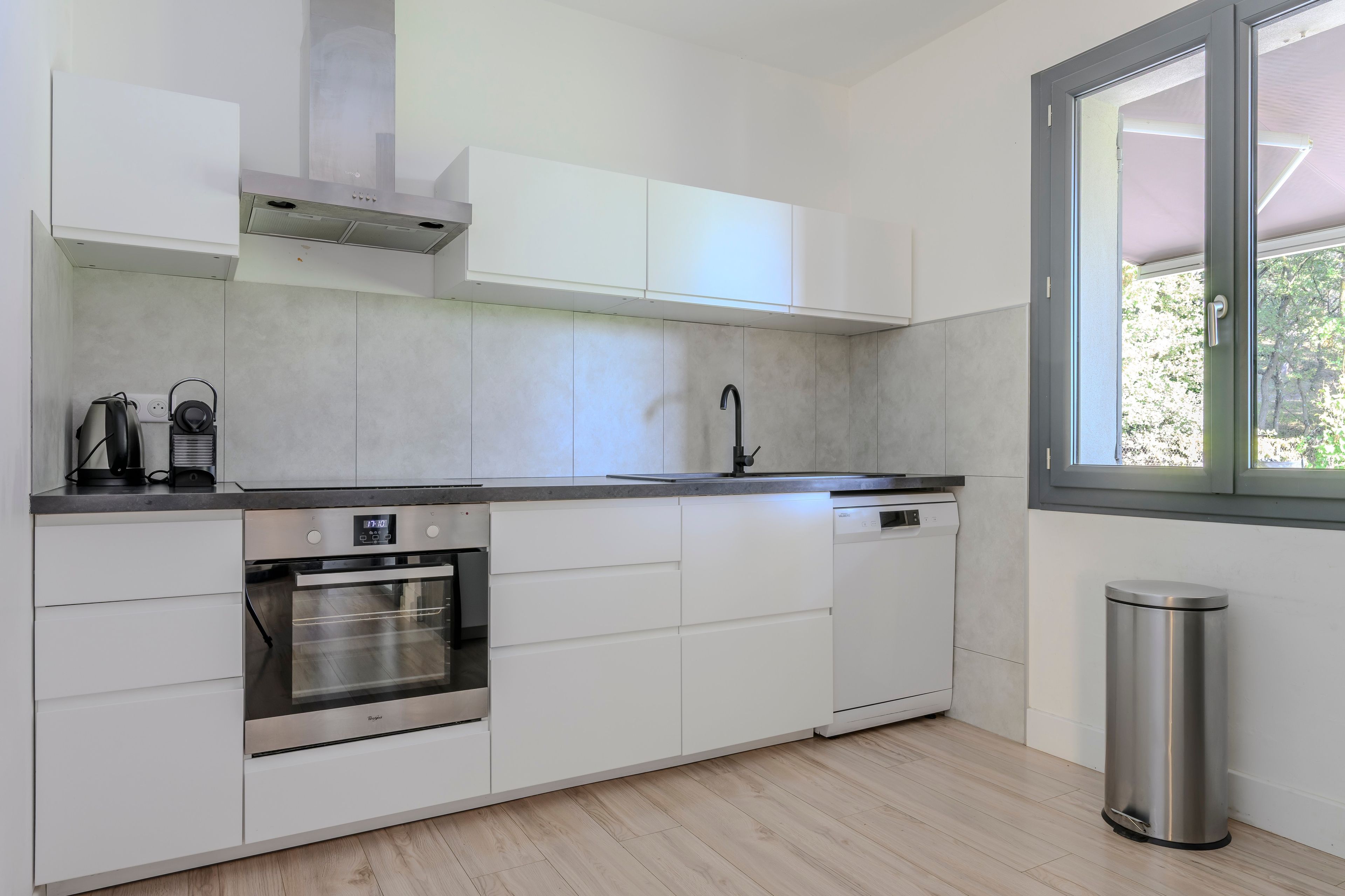 A modern kitchen with white cabinetry, a stainless steel oven, a dishwasher, and a window providing natural light.