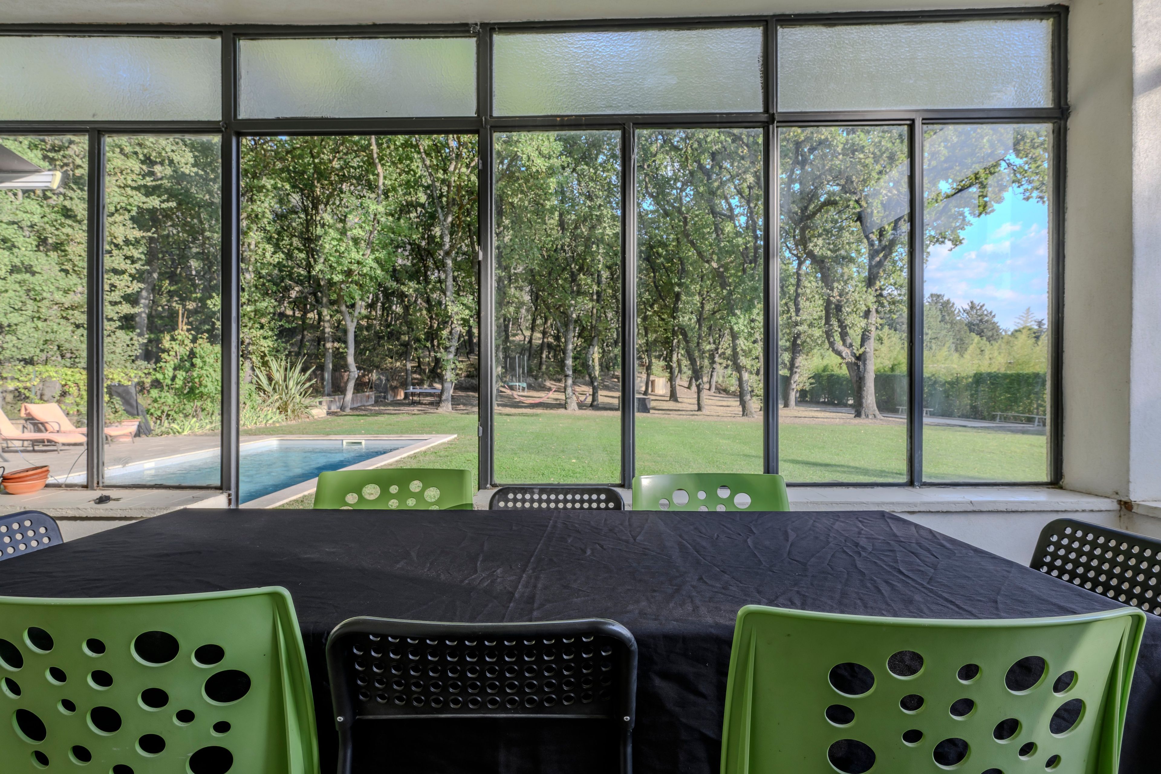 Une pièce dotée d’une grande fenêtre donnant sur une piscine et un espace verdoyant avec des arbres. On y trouve une table recouverte d’une nappe noire et des chaises vertes.